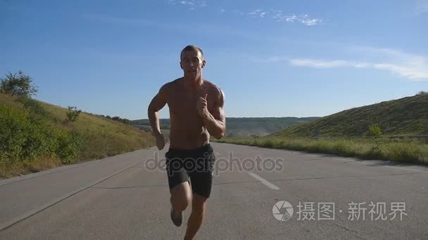 在公路慢跑的短跑运动员男人。男运动员在夏季训练户外。年轻强壮肌肉的人在锻炼期间在农村公路上行使。外积极健康的生活方式。慢动作