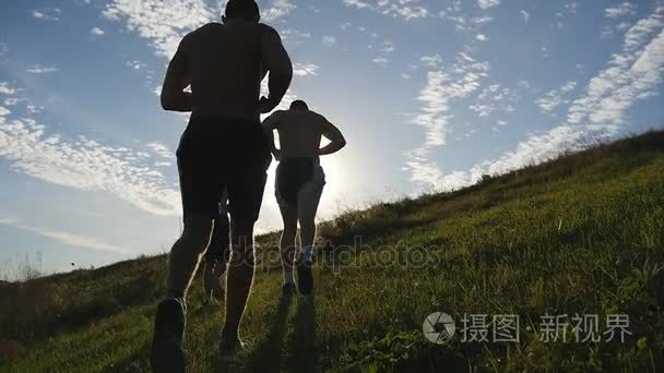 年轻男子在蓝色天空背景下运行过绿色的小山。日落时分，男运动员慢跑性质。体育跑步慢跑上山室外日出与耀斑。越野训练。慢动作