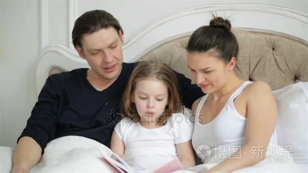 漂亮的小女孩阅读坚持书躺在床上和她的父母。帅父是亲吻他聪明的孩子