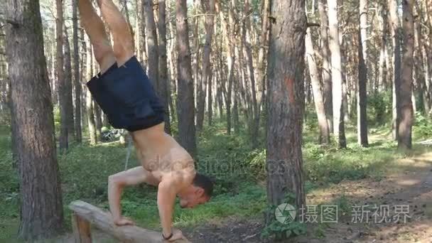 强大的肌肉男做倒立在森林里。肌肉发达的男性健身家伙做日志在木头上的特技。运动员训练执行倒立在外肌。健身运动健康的生活方式