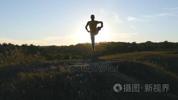 轮廓的运动的人站在室外的瑜伽姿势。瑜伽练习瑜伽移动，并在自然中的位置。运动员在一条腿上保持平衡。美丽的天空和太阳作为背景。健康积