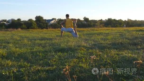 年轻男子坐在绿草的草地，做瑜伽练习。肌肉发达的家伙在自然伸展他的身体。在晴朗的一天训练户外。在背景的美丽景观。关闭视频