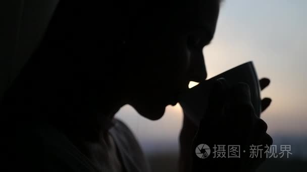 配置文件的一个美丽的女孩与一杯茶在手反对窗外美丽的日落。1920 x 1080。 慢动作