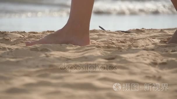 女性的脚走在金色的沙滩，在沙滩与海洋波浪在背景的特写。双腿踩在沙子的年轻女子。在海边的赤脚女孩。夏季放假的日子。慢动作