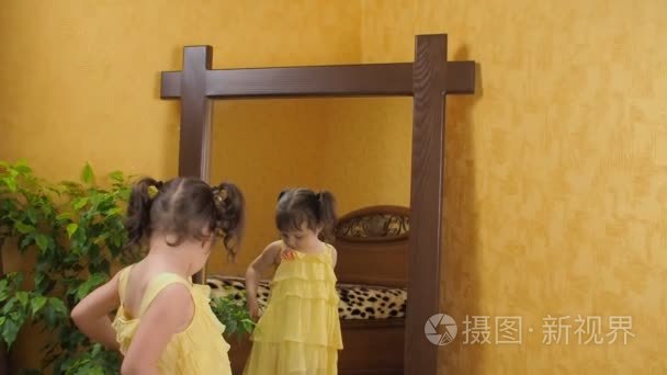 一个小女孩看着镜子。一个漂亮的女孩，在她头上的尾巴抱着她的衣服。一个孩子在一件黄色的连衣裙的镜子