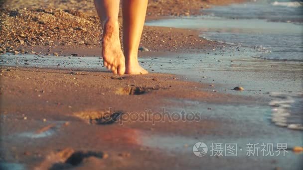 女性美丽晒黑的双腿走在海滩上的沙子。女孩在日落时分海边散步