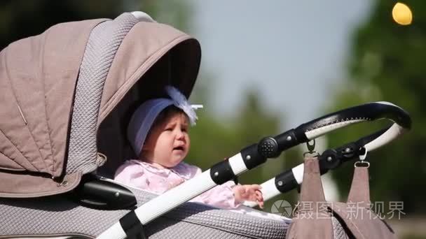 哭宝贝女孩坐在婴儿车在公园里在阳光的一天