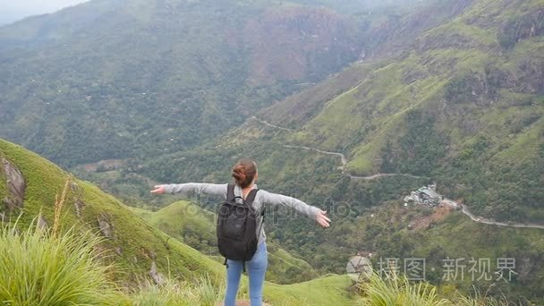 年轻的女性的徒步旅行者背包达山和举的手的顶部。女游客站在美丽的峡谷的边缘，凯旋扩张军备了。慢动作后方回来查看