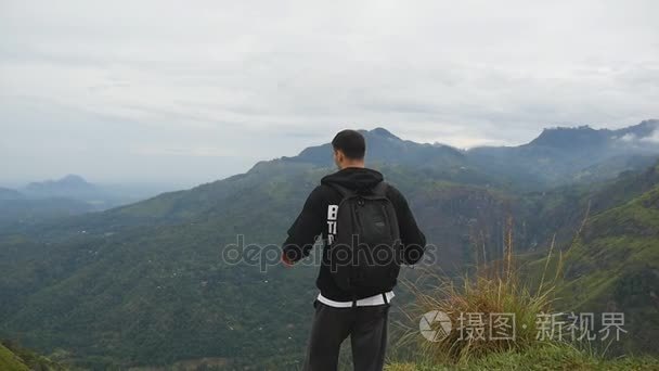 年轻的男性的徒步旅行者背包达山和举的手的顶部。男子旅游站在美丽的峡谷的边缘，凯旋扩张军备了。慢动作后方回来查看