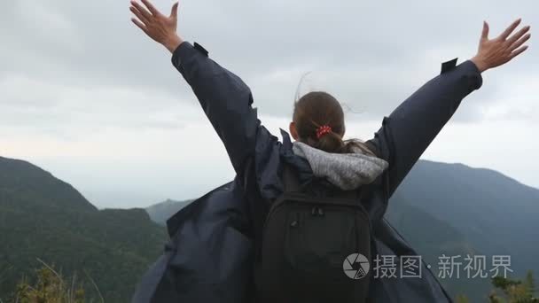 女游客站在美丽的峡谷的边缘，凯旋扩张军备了。年轻的女性的徒步旅行者背包达山和举的手的顶部。慢动作后方回来查看