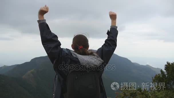 女游客站在美丽的峡谷的边缘，凯旋扩张军备了。年轻的女性的徒步旅行者背包达山和举的手的顶部。慢动作后方回来查看