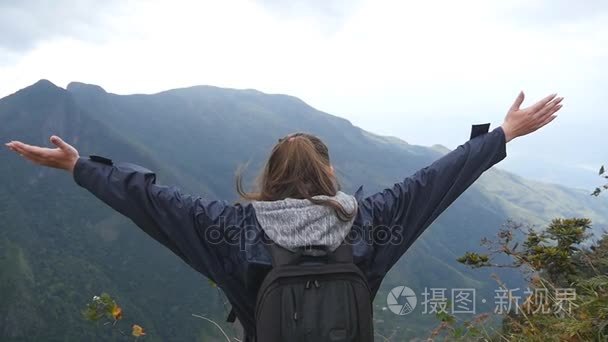 年轻的女性的徒步旅行者背包达山和举的手的顶部。女游客在雨衣站在边缘，美丽的峡谷，凯旋扩张军备了。后方的背影