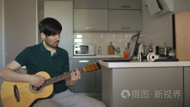 有魅力的年轻人坐在厨房学弹吉他在家里使用便携式计算机