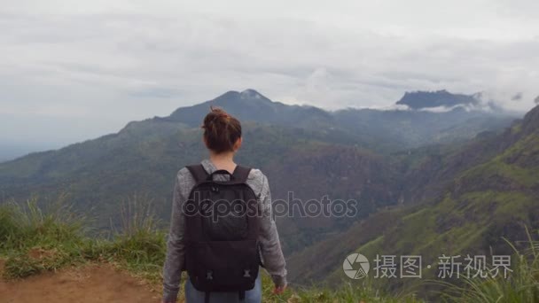 年轻的女性的徒步旅行者背包达山和举的手的顶部。女游客站在美丽的峡谷的边缘，凯旋扩张军备了。后方的背影