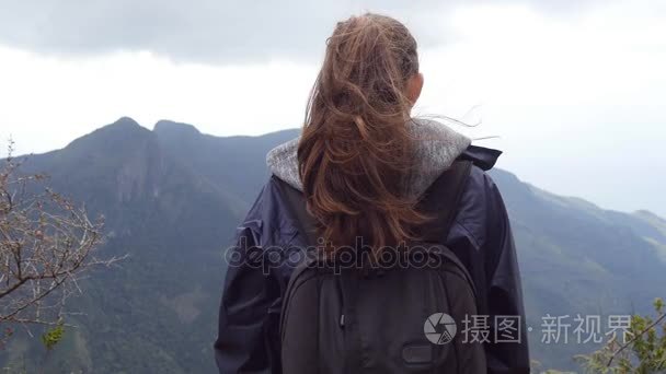 变得面目全非的女游客站在美丽的峡谷的边缘上，欣赏风景。年轻的女性达到顶山和举的手上，背包里的雨衣徒步旅行者。后方的背影