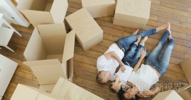 顶视图的英俊的父亲和母亲玩和拥抱他们的小儿子躺在地板上。家庭放松和收拾好行李搬家，从纸箱后笑查看从顶部