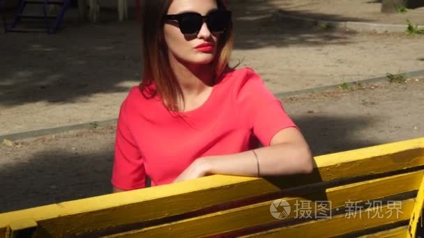 太阳镜和红色西装位漂亮的女士坐在黄色的长凳上，公园
