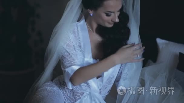 年轻的吸引力新娘在优雅的白色内衣，坐在床上闲荡。美丽的女人，与摆在卧室的面纱