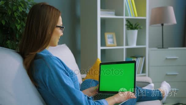 年轻的女人坐在客厅沙发上用绿屏的笔记本电脑。女人手在笔记本电脑键盘上打字。色度键。背面的看法