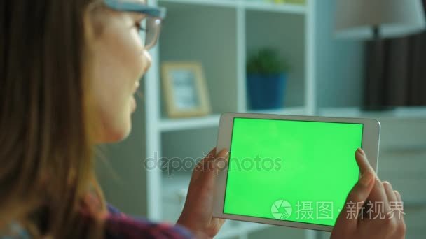 年轻的女人坐在客厅沙发上用绿屏白色平板电脑。女人手滚动 滑动页面。色度键。背面的看法。关闭