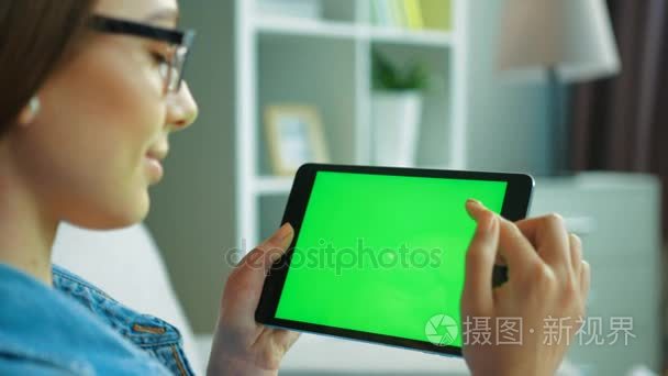 坐在客厅沙发上用绿屏黑色平板电脑年轻漂亮的女人。女人手滚动、 缩放页面。色度键。查看在肩上。关闭