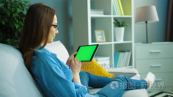 轻松年轻女人使用黑色平板电脑与绿屏坐，躺在客厅沙发上。女人手滚动、 缩放页面。色度键。侧视图