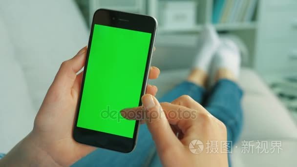 黑色的智能手机用绿屏的年轻女人。手持移动电话在垂直的位置躺在客厅沙发上的女人。色度键。关闭。顶视图