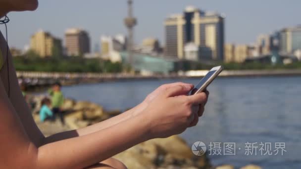 使用智能手机坐在路堤附近的海和大城市的摩天大楼上的年轻女人视频