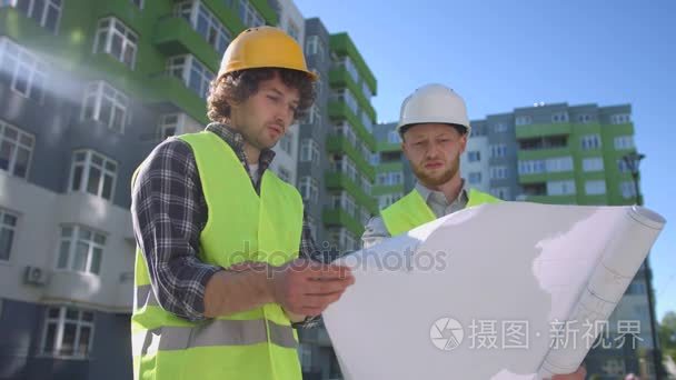 两个年轻的建筑师在防护头盔和绿色安全夹克，讨论，看图纸上。户外