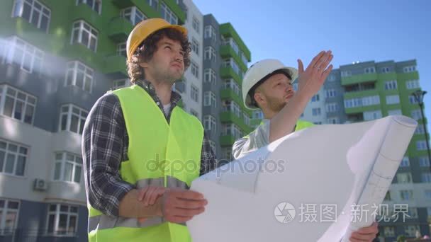 两位白种人建筑师在防护头盔和绿色安全夹克，讨论，看图纸和施工过程。户外