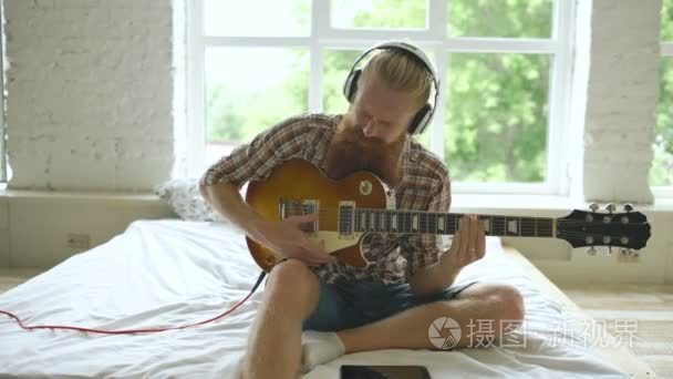 有吸引力的大胡子的男人，坐在床上学习弹吉他在现代居室在家中使用平板电脑