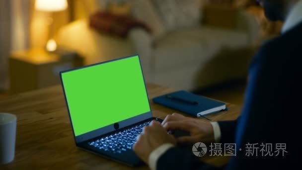 在家里的男人坐在他的办公桌和便携式计算机上与绿色屏幕上它的类型。他的公寓是在黄色色彩和温暖