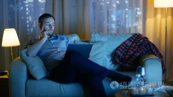 在晚上中间岁的男子坐在沙发上持有笔记本电脑上在他的膝盖和 It.Tv 的工作是和它照亮 Him.Skyscrapers 从窗口看视频