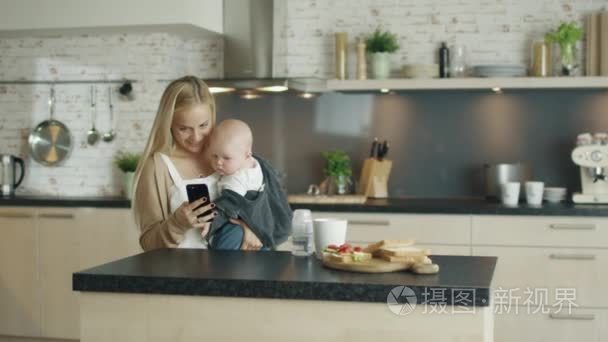 年轻的母亲认为她美丽的孩子却不能在厨房。她也用她的智能手机用一只手，孩子看起来很感兴趣视频