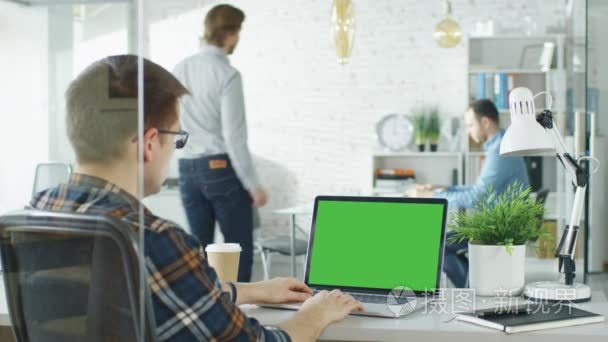 一个人坐在办公桌前与绿色屏幕笔记本电脑的特写镜头。背景模糊和明亮亮的办公室，两个年轻男子握手，坐下来的谈话