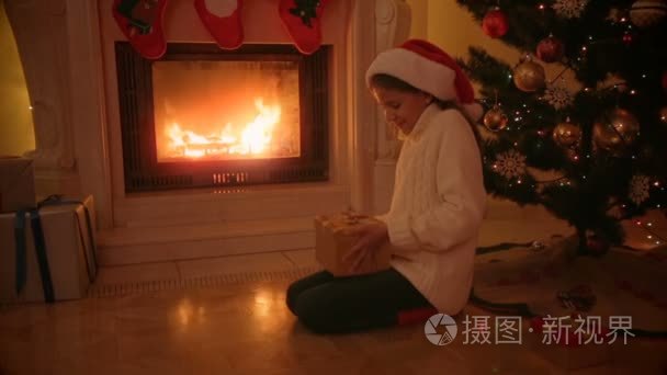 快乐的女孩，在圣诞老人的帽子，坐在火炉边，看里面发光的礼物盒