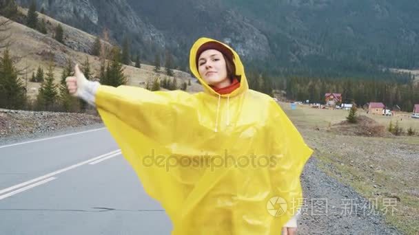 旅行者女人多云路上搭便车和行走。年轻快乐的背包客女人寻找搭车，开始一段旅程。穿着黄色的雨衣，雨中