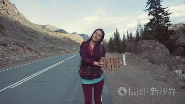 十几岁的女孩举冒险牌子上山路 微笑和大笑。搭便车去历险