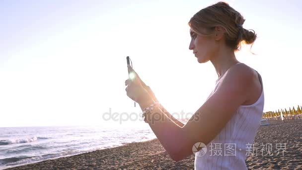 漂亮的金发女人采取自拍照与智能电话海滩上的日落