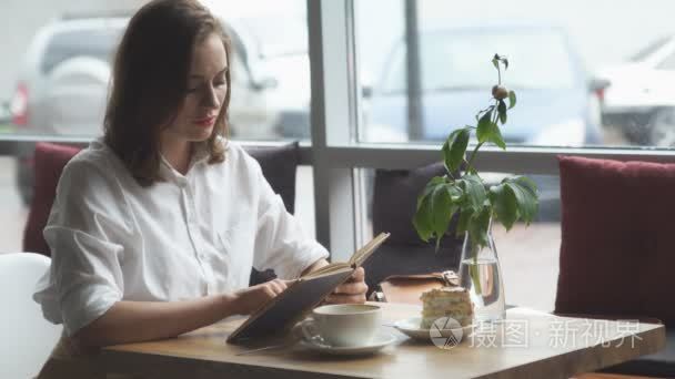 那个女孩正在读一本书，坐在一个咖啡馆和享受咖啡和蛋糕