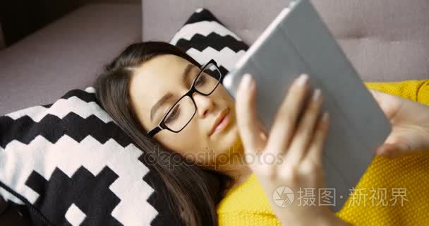 在玻璃上平板电脑躺在客厅的沙发上在线读书的年轻女子