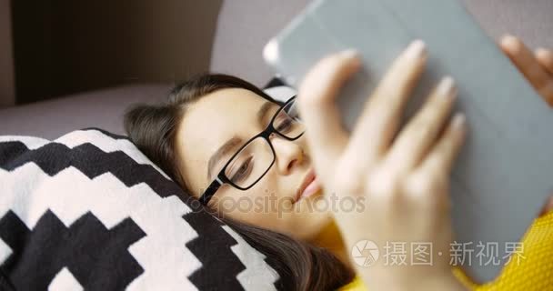 有吸引力的年轻女子躺在沙发上使用平板电脑设备。美丽的女人在平板电脑和微笑在家里上打字的眼镜。关闭