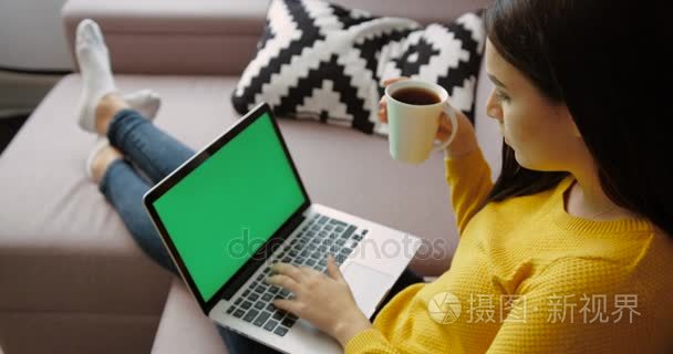 美丽的年轻女子笔记本 pc 使用绿屏，坐在客厅沙发上喝咖啡。后面的观点。色度键