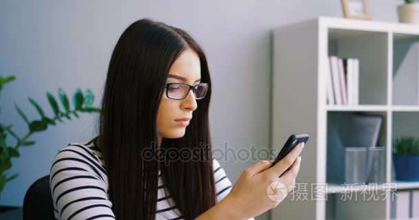 迷人的女人在办公室使用智能手机的眼镜。关门时坐在办公室桌前在室内使用手机的女商人。近摄镜头。拍摄的肖像