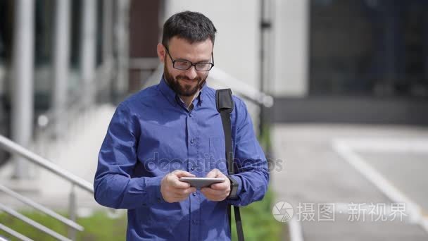英俊的大胡子的男人穿着蓝衬衫和棕色的裤子是站在户外和打字使用他的智能手机的消息。年轻的商人正在等待朋友在街上晴朗的一天