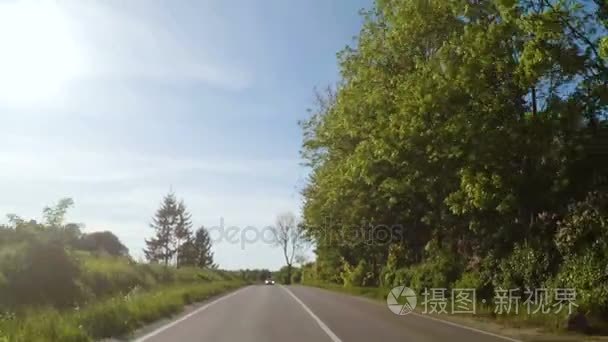 乘汽车旅行圆的欧洲。装的摄像头，后面的观点。驾驶对长直柏油路向太阳在欧洲的相机视图。在路边的两车道公路上的树木。耀眼的太阳升起时