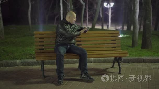 在春天，秃头的男人坐在公园的长椅上，看着他的手机在路灯的夜晚