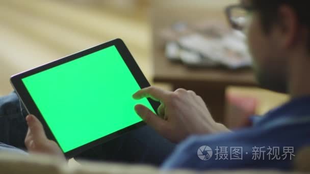 男人放在家里的沙发上和绿色屏幕在横向模式中使用平板电脑