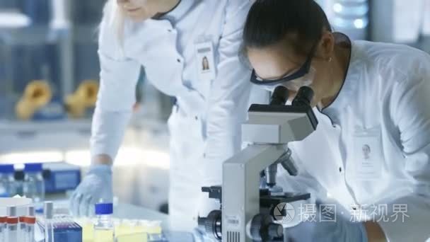 医学研究的科学家滴样本幻灯片和她的同事检查镜下。他们在一个现代的实验室工作