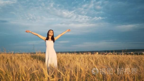 一个年轻的姑娘站在金色的田野的小麦在日落时分和引发他举起手来，慢动作